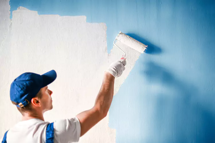 Painter using a roller to apply white paint to a blue wall
